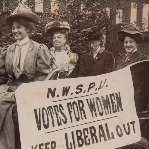A group of four suffragettes in a car holding a sign that reads ‘Votes for Women, Keep Liberal Out’