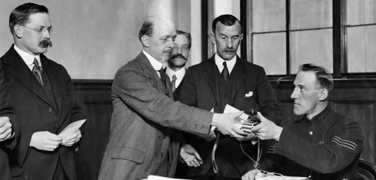 Black and white photo showing a man seated at a desk in uniform, handing a man standing some papers.
